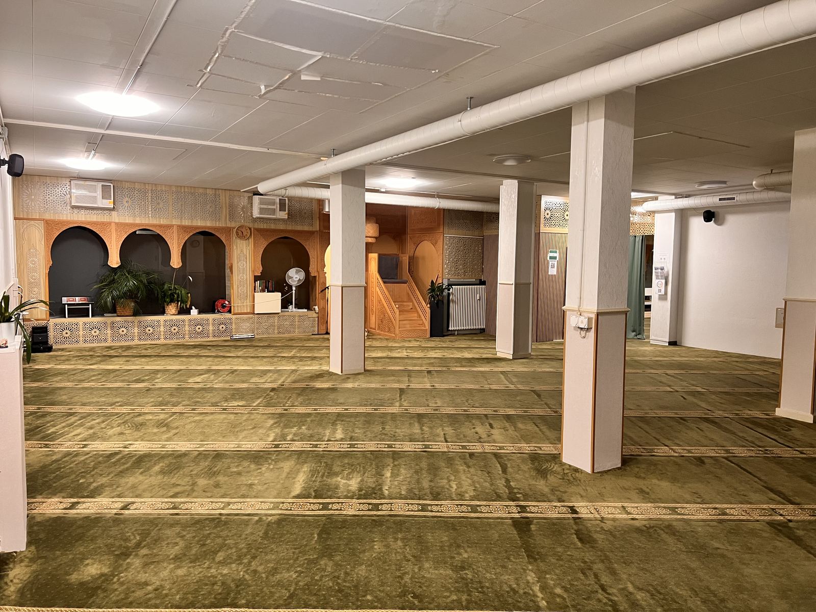 Weiter Gebetsraum mit grünem Teppich, hellen Säulen und einer Wand aus geschnitzten Holzbögen im Hintergrund.