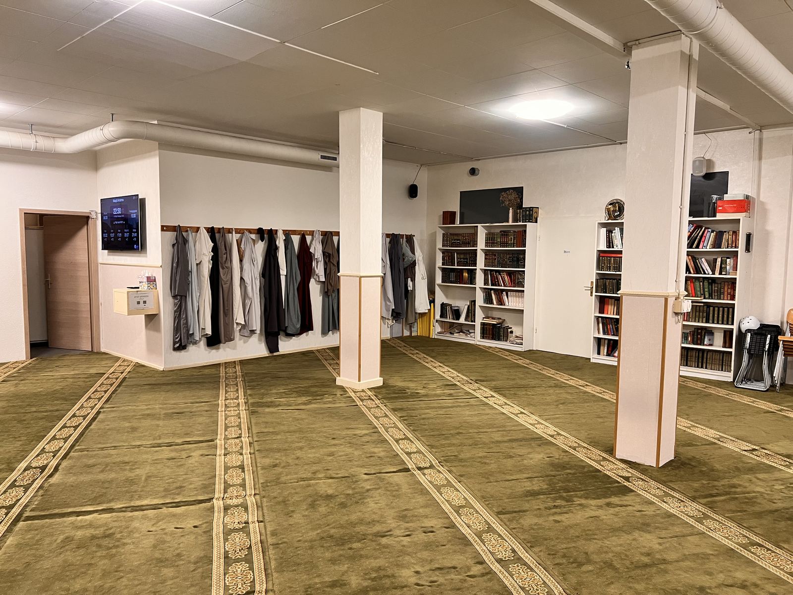 Seitenwand des Gebetsraums mit einer Reihe aufgehängter Jacken und weissen Bücherregalen.