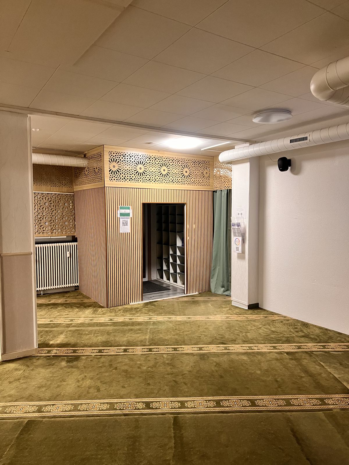 Holzverkleideter Durchgang mit Ornamentgitter darüber, dahinter offene Schuhfächer.