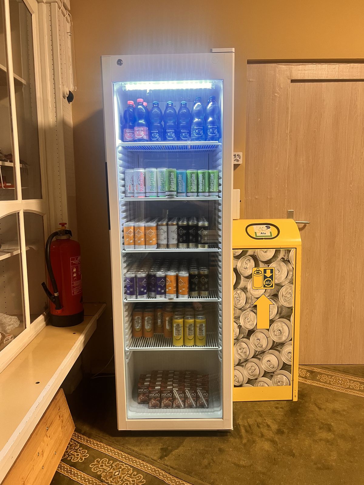 Beleuchteter Glaskühlschrank mit Getränkeflaschen und Dosen, daneben ein gelber Becherspender.